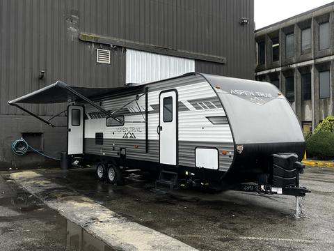 A 2022 Aspen Trail travel trailer with a sleek gray exterior features a pull-out awning and is parked next to a building with a hose connected to it
