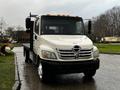 A 2006 Hino 155 truck with a flatbed and a chrome front grille parked on a wet surface displaying its large wheels and distinctive front design