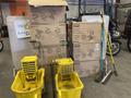 Ten yellow mop buckets and assorted brooms in various colors stacked beside each other