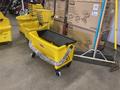 Ten yellow mop buckets and assorted brooms are stacked and arranged with a focus on a yellow mop bucket with a wringer in the foreground