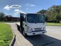 A white 2015 Isuzu NPR truck with a flatbed and large cab parked on a street with trees in the background