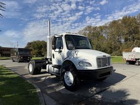 A white 2003 Freightliner M2 106 truck with a silver exhaust pipe and large tires parked on the street