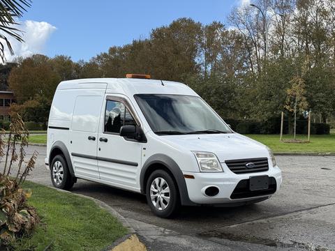 A white 2012 Ford Transit van with a high roof and orange warning light on top parked on a street with trees in the background