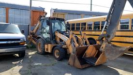 A 2015 Case 580 backhoe loader with a front bucket and rear excavator arm parked between a white van and yellow school buses