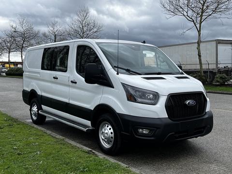 A 2023 Ford Transit van in white with a black grille and gray accents parked on grass with a cloudy sky in the background