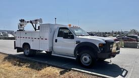 A white 2008 Ford F-550 utility truck with a mounted crane and storage compartments on the side parked on a roadside