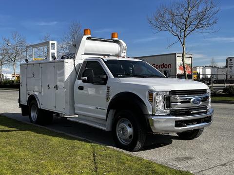 A 2018 Ford F-550 utility truck with a white exterior features a ladder rack and storage compartments on the side and an orange light bar on the roof