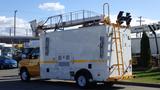 A 2015 Ford Econoline work truck with a customized white and yellow exterior featuring a raised light bar and a ladder on the side for access to the roof