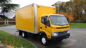 A 2009 Hino 145 truck with a yellow boxy cargo area and a cab featuring a black bumper and front grille, parked with its side facing the viewer