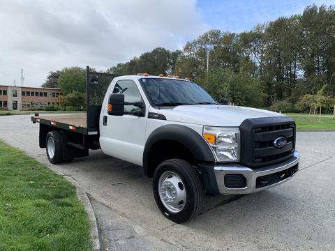 A 2012 Ford F-550 flatbed truck with a white cab and black front grille parked on a surface road