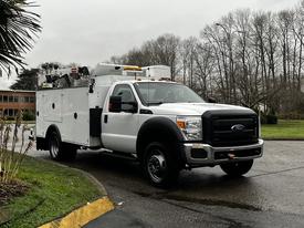 A 2015 Ford F-550 utility truck with a white exterior and an equipment compartment in the back parked on a road