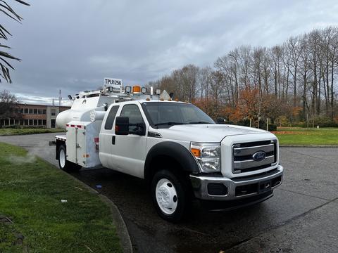 A 2016 Ford F-550 white pickup truck with a tank on the back and warning lights on top parked on a roadway