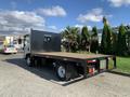 A 2015 Isuzu NPR truck with a flatbed and a sturdy black rear guard parked on a paved surface