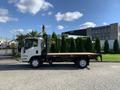 A white 2015 Isuzu NPR flatbed truck parked on a driveway with a flat wooden bed and a cab designed for transporting goods