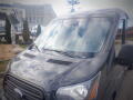 A 2015 Ford Transit van with a gray exterior and a front windshield displaying reflections of clouds